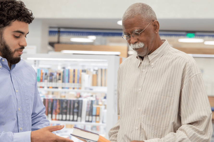 A senior man in a library engaged in a conversation with a younger man while holding a book.