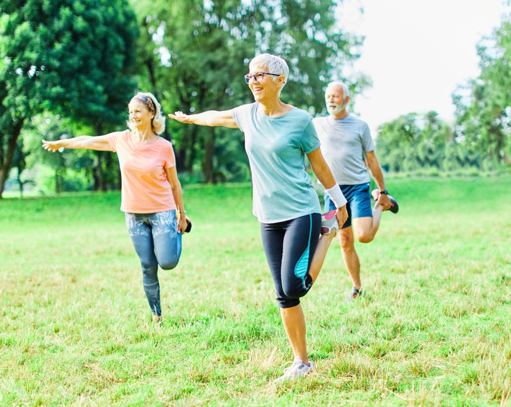 A group of active seniors stretching and exercising outdoors in a lush green park. They are smiling and engaging in a fitness routine, emphasizing health and wellness in a retirement community.